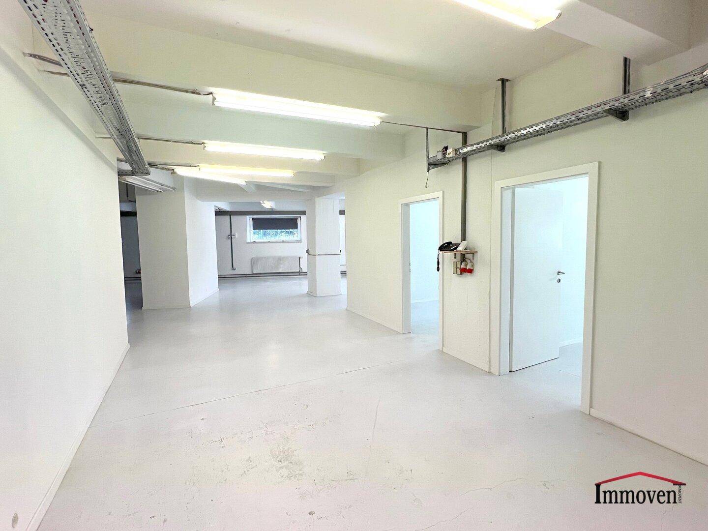 Heller, offener Gewerberaum mit weißen Wänden, Betonboden und Zugang zu weiteren Räumen.