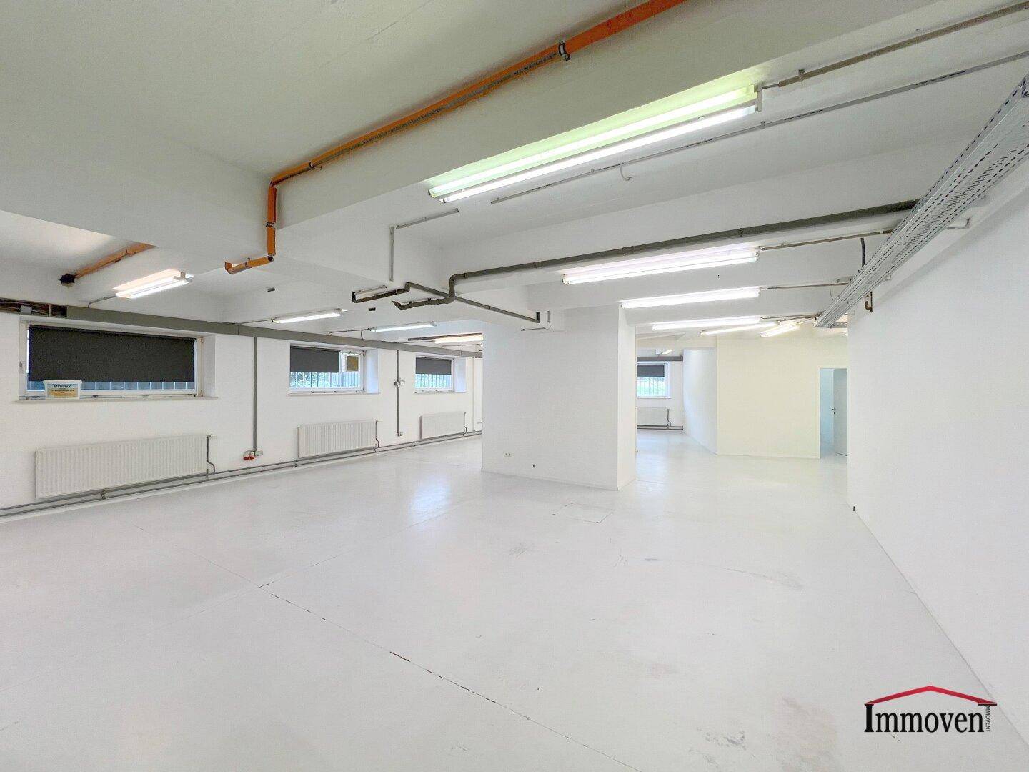 Weitläufiger, heller Gewerberaum mit sichtbaren Installationen an der Decke und mehreren Fenstern.