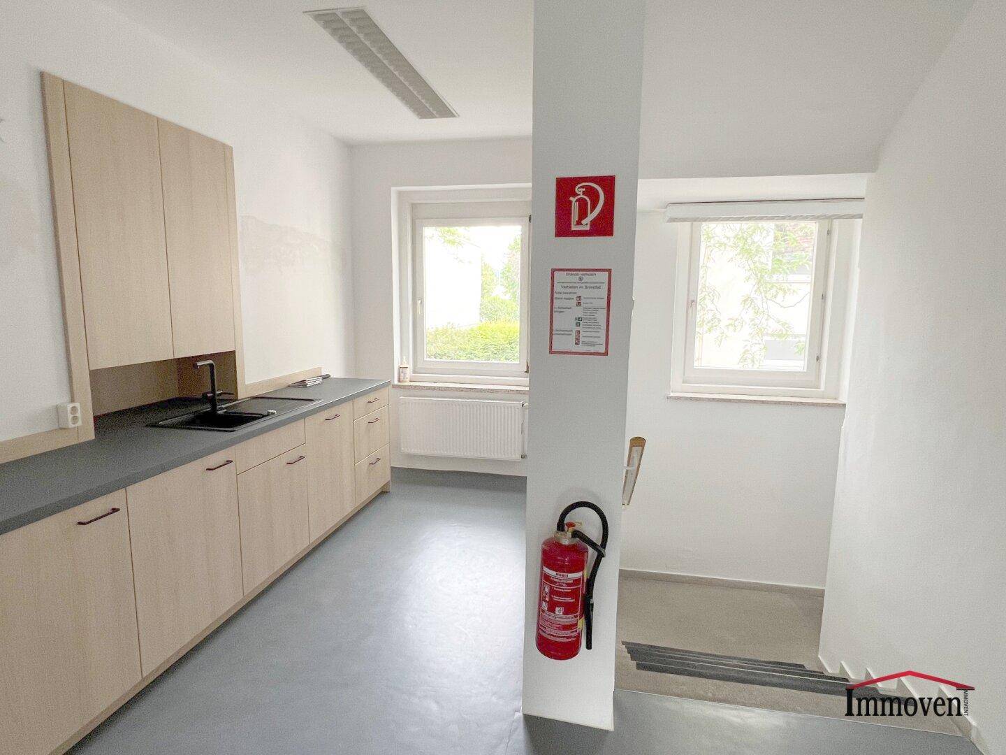 Funktionale Küchenzeile mit hellen Holzschränken, Spüle und Fenstern, daneben ein Treppenaufgang.