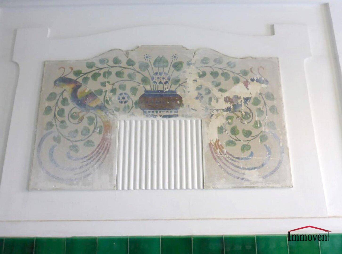 Detail einer Wanddekoration mit floralen Mustern und Vogelmotiven im historischen Stil über grünen Fliesen.