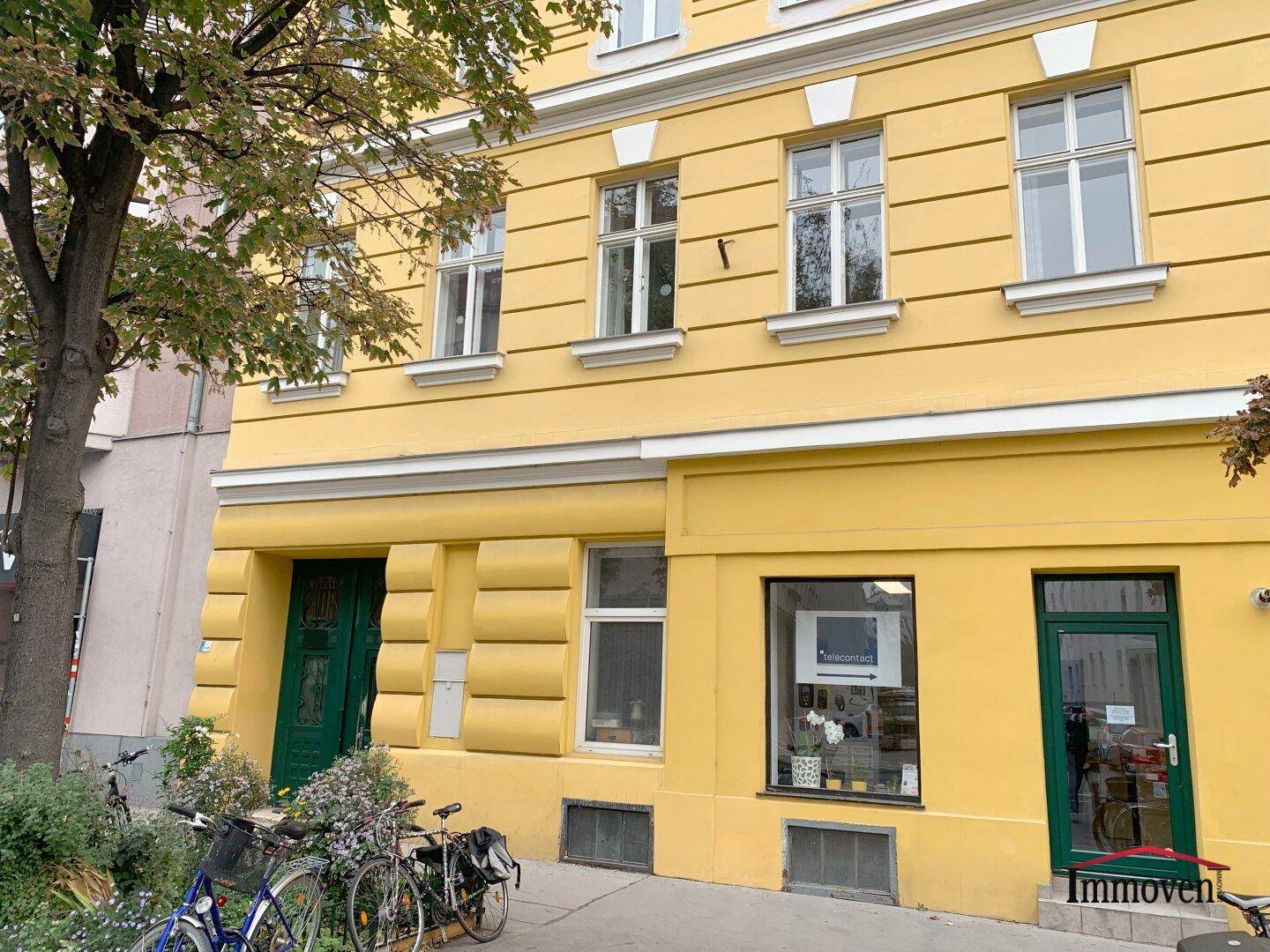 Erdgeschoss eines gelben Gebäudes mit grünen Türen, Schaufenstern und Fahrrädern auf dem Bürgersteig.