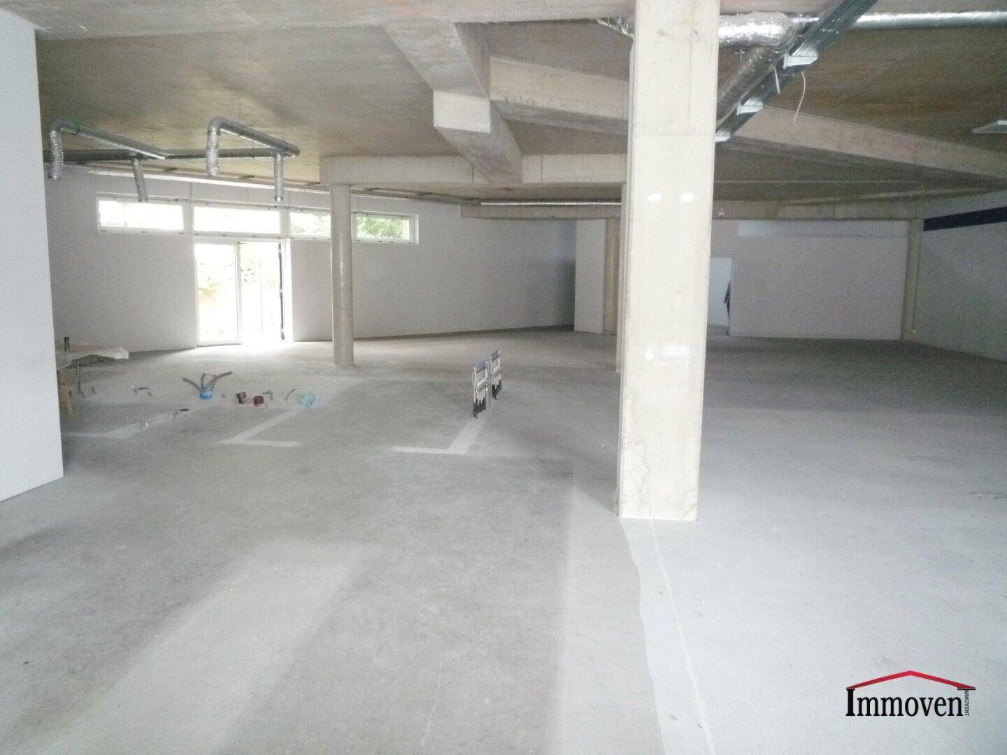 Heller Gewerberaum im Rohbau mit großen Fenstern, Betonsäulen und sichtbaren Installationen und grünem Ausblick.