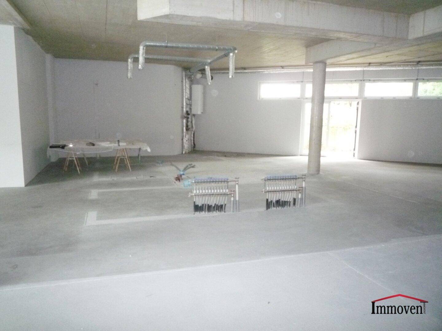 Unfertiger Gewerberaum mit Betonboden, sichtbaren Rohren und einem provisorischen Arbeitstisch im Rohbauzustand.