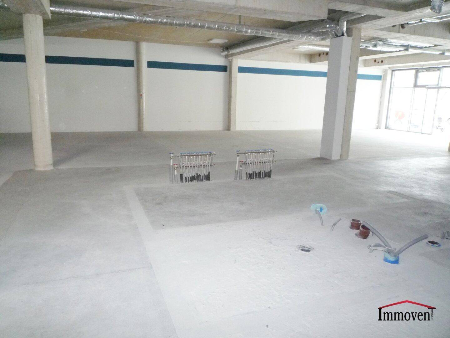 Großer Gewerberaum mit Betonboden, blauen Wandstreifen und vorbereiteten Bodeninstallationen im Rohbauzustand.