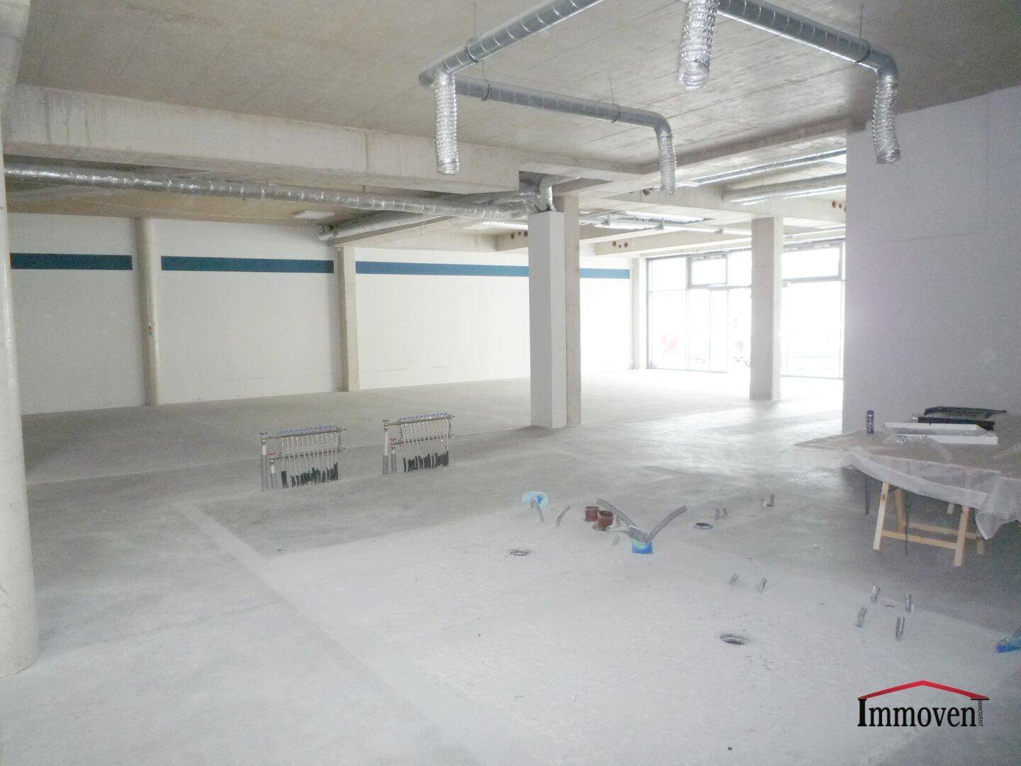 Heller Gewerberaum mit Betonboden, sichtbaren Lüftungsrohren und großen Fensterfronten im Rohbauzustand.