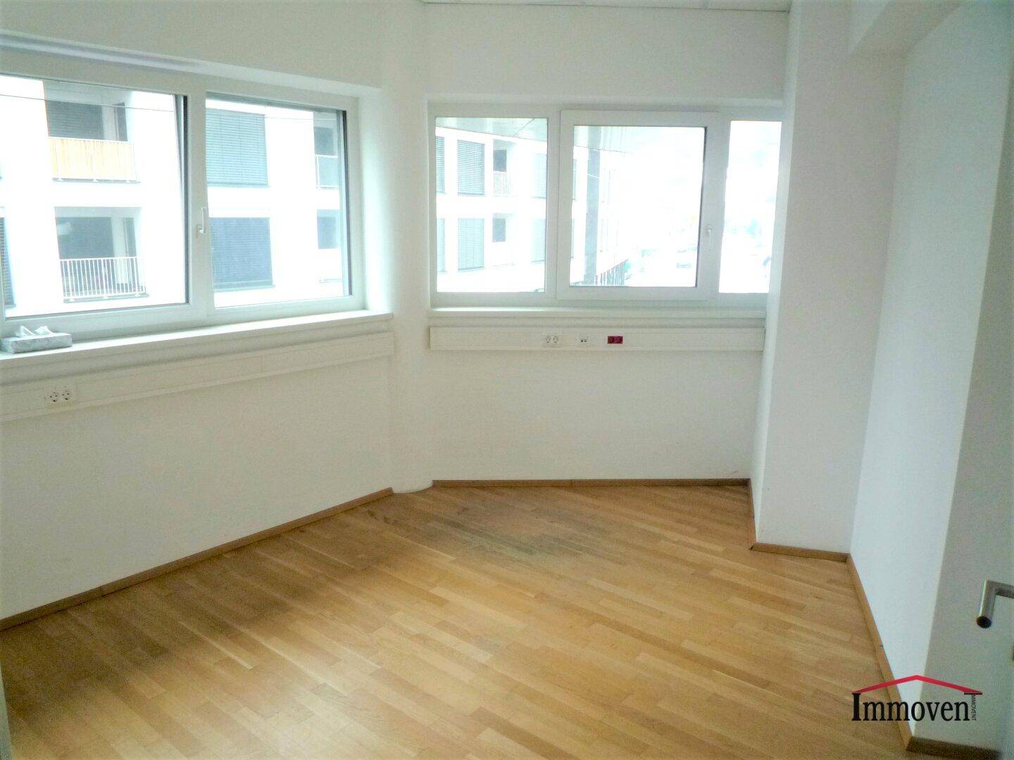 Leerer Büroraum mit Holzboden und mehreren Fenstern, die auf andere Gebäude blicken.