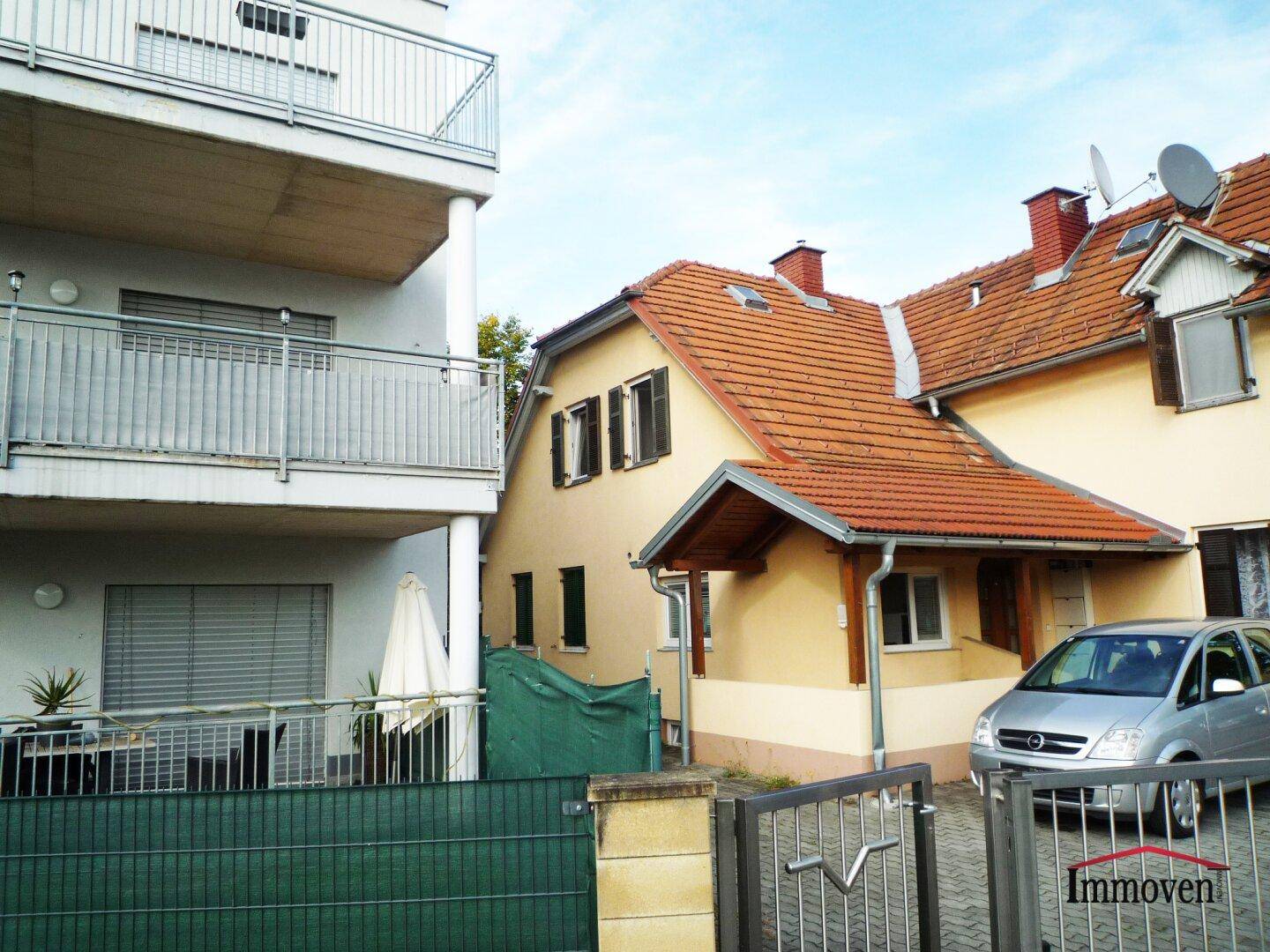 Seitenansicht des Hauses mit Satteldach und angrenzendem modernen Apartmentgebäude mit Balkonen.