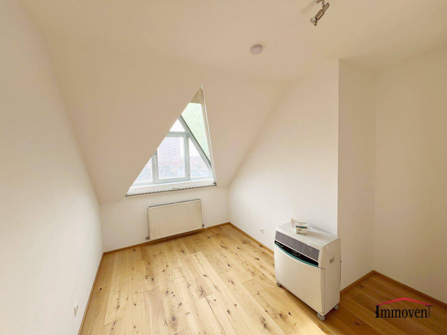 Dachzimmer mit Holzboden, dreieckigem Fenster und einer mobilen Klimaanlage für Komfort.