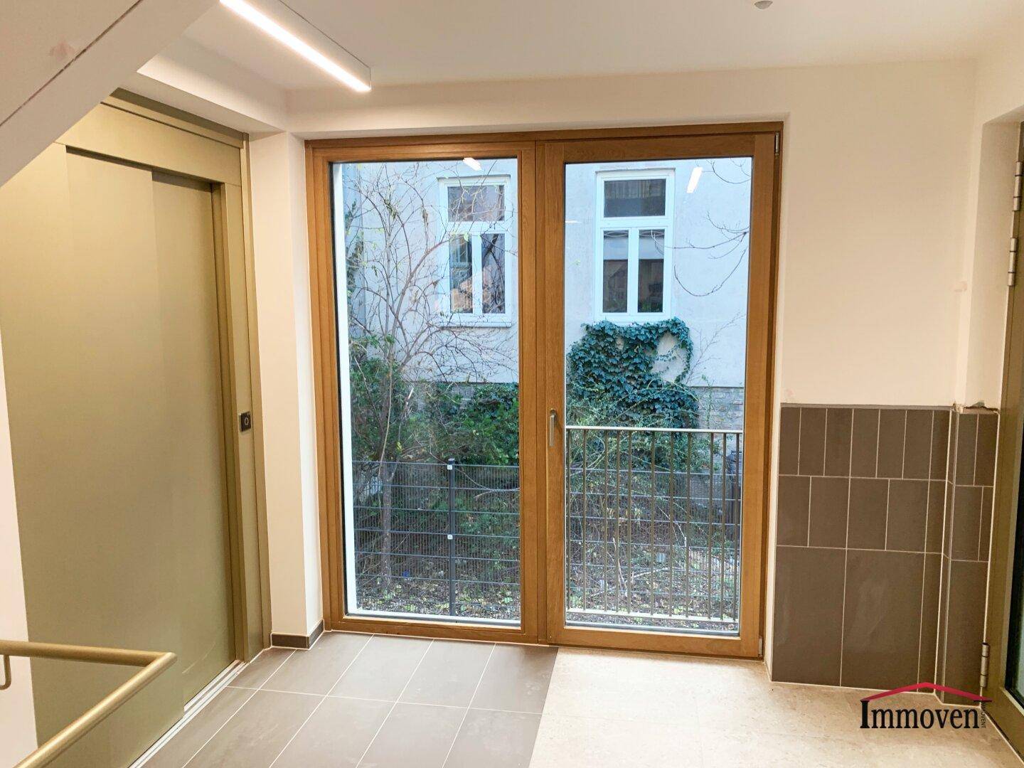 Heller Eingangsbereich mit Aufzug, großen Fenstern und Blick auf einen begrünten Innenhof.