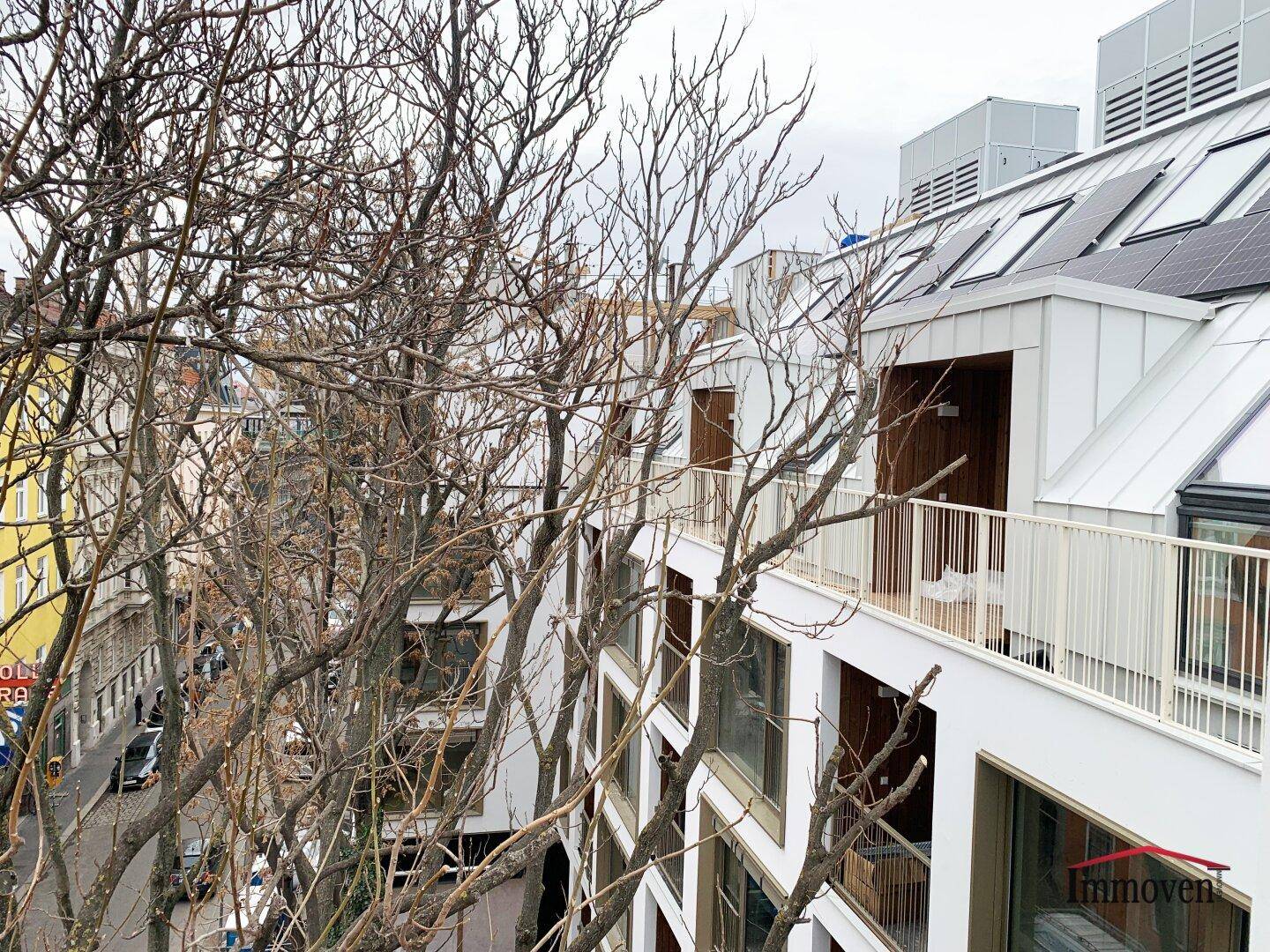 Blick von einem Balkon durch kahle Äste auf eine belebte Straße und umliegende Gebäude.