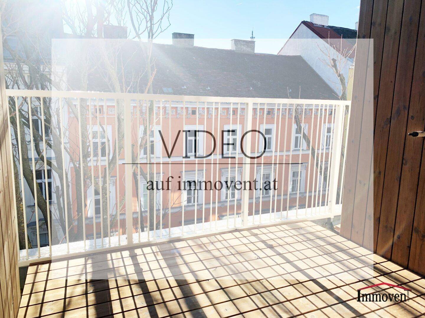 Heller Balkon mit Holzfußboden und weißem Geländer, bietet einen Blick auf gegenüberliegende Gebäude.