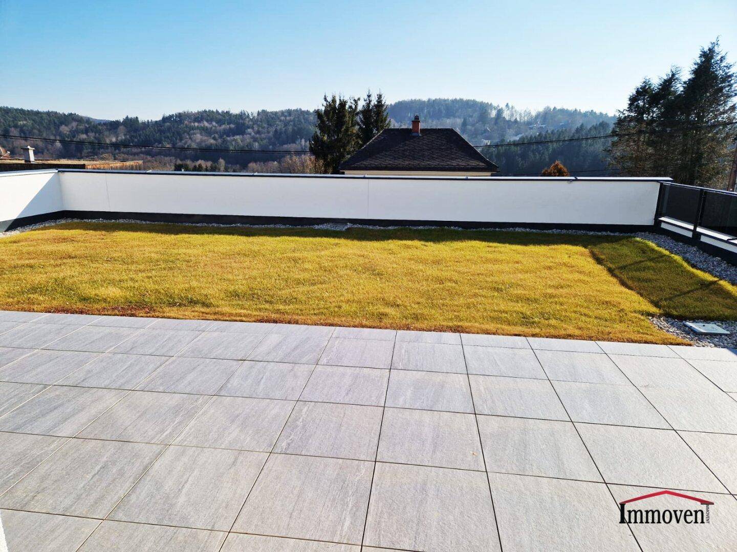 Geräumige Terrasse mit Fliesenboden und angrenzender Rasenfläche, umgeben von einer niedrigen Mauer.
