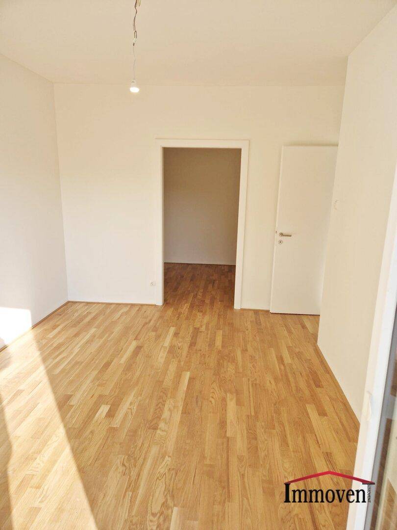 Unmöblierter Raum mit hellem Parkettboden und einer Tür, die zu einem weiteren Zimmer führt.