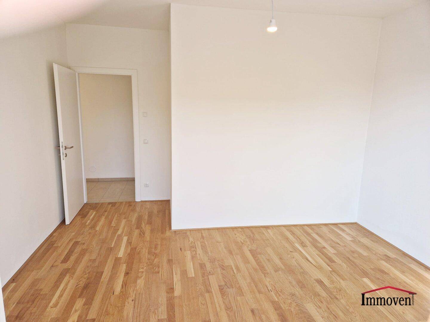 Unmöblierter Raum mit Parkettboden und einer offenen Tür, die in einen angrenzenden Bereich führt.