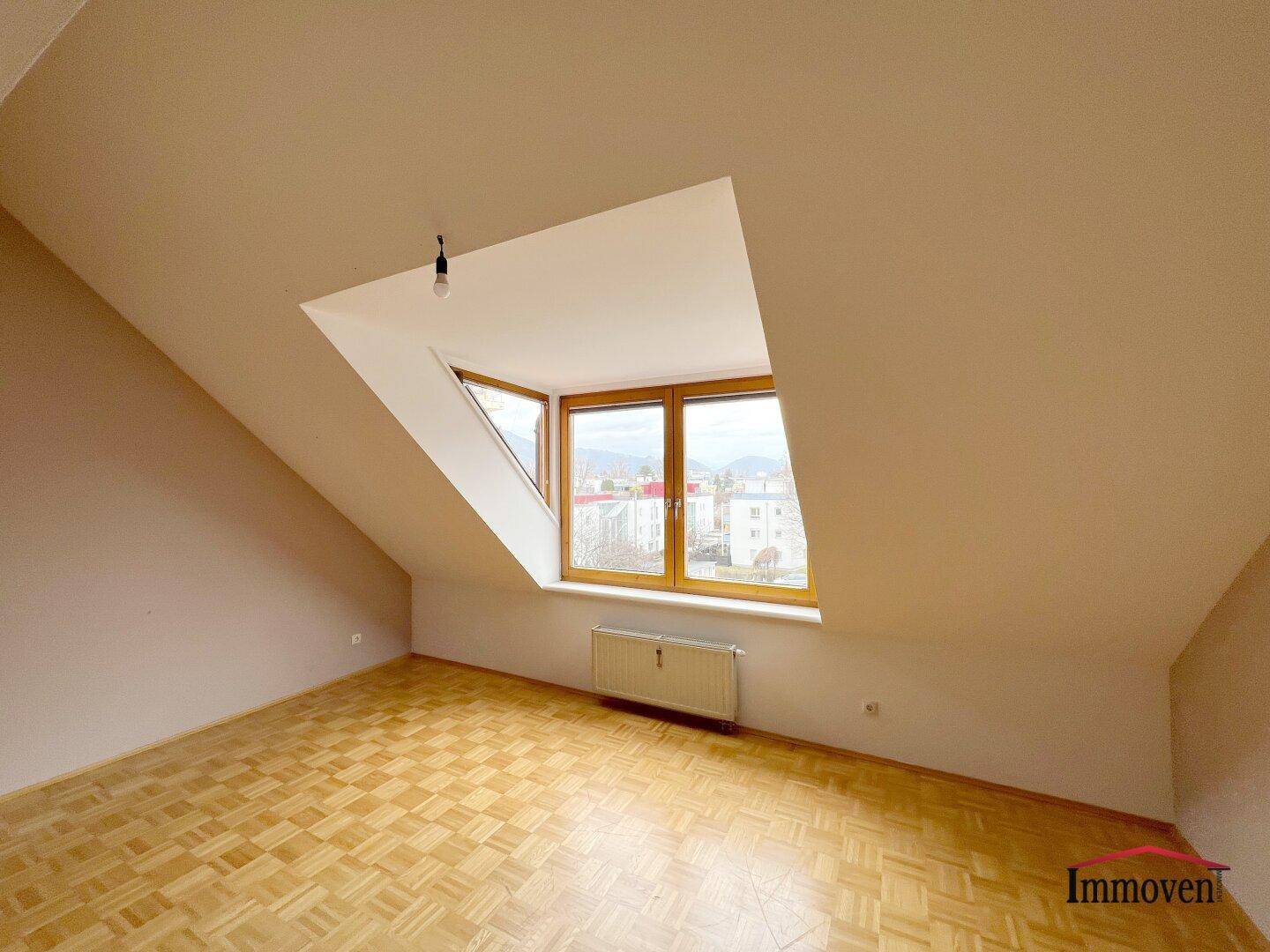 Helles Dachgeschosszimmer mit Parkettboden, schrägen Wänden und einem großen Fenster mit Ausblick.