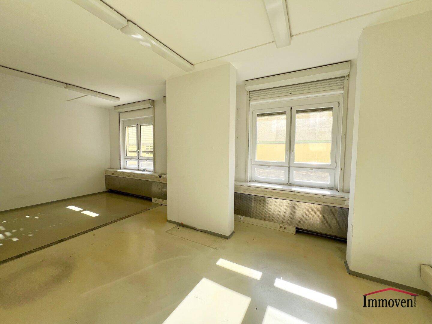 Heller Büroraum mit zwei Fenstern, Jalousien und Heizkörpern unter den Fensterbänken.