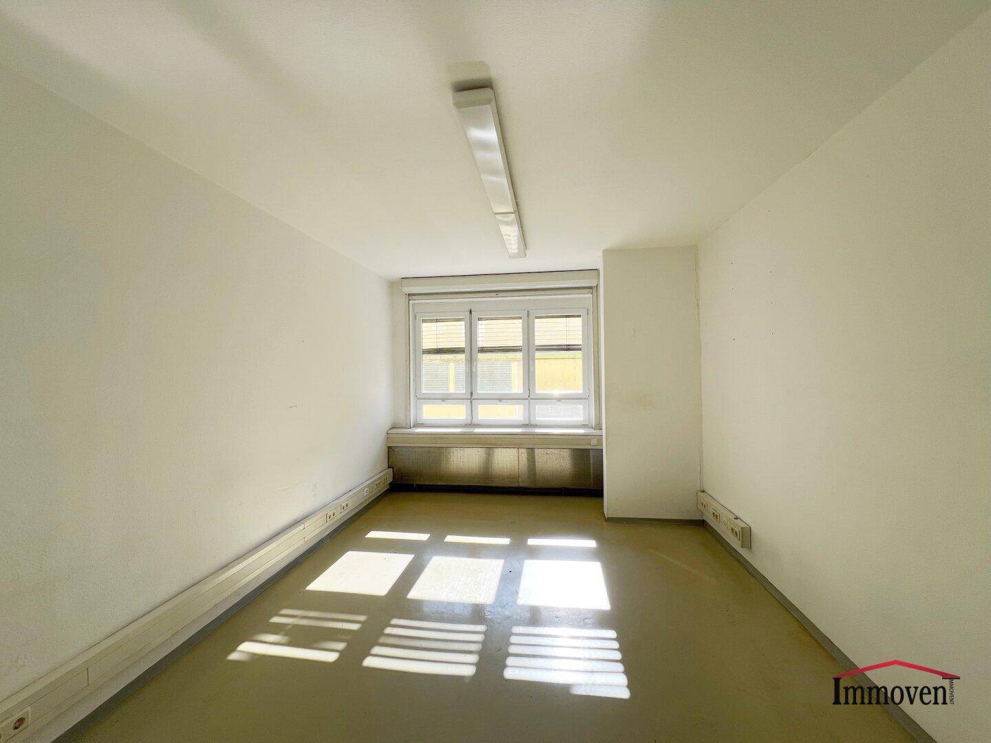 Leerer Büroraum mit einem großen Fenster, Jalousien und Streifen von Sonnenlicht auf dem Boden.