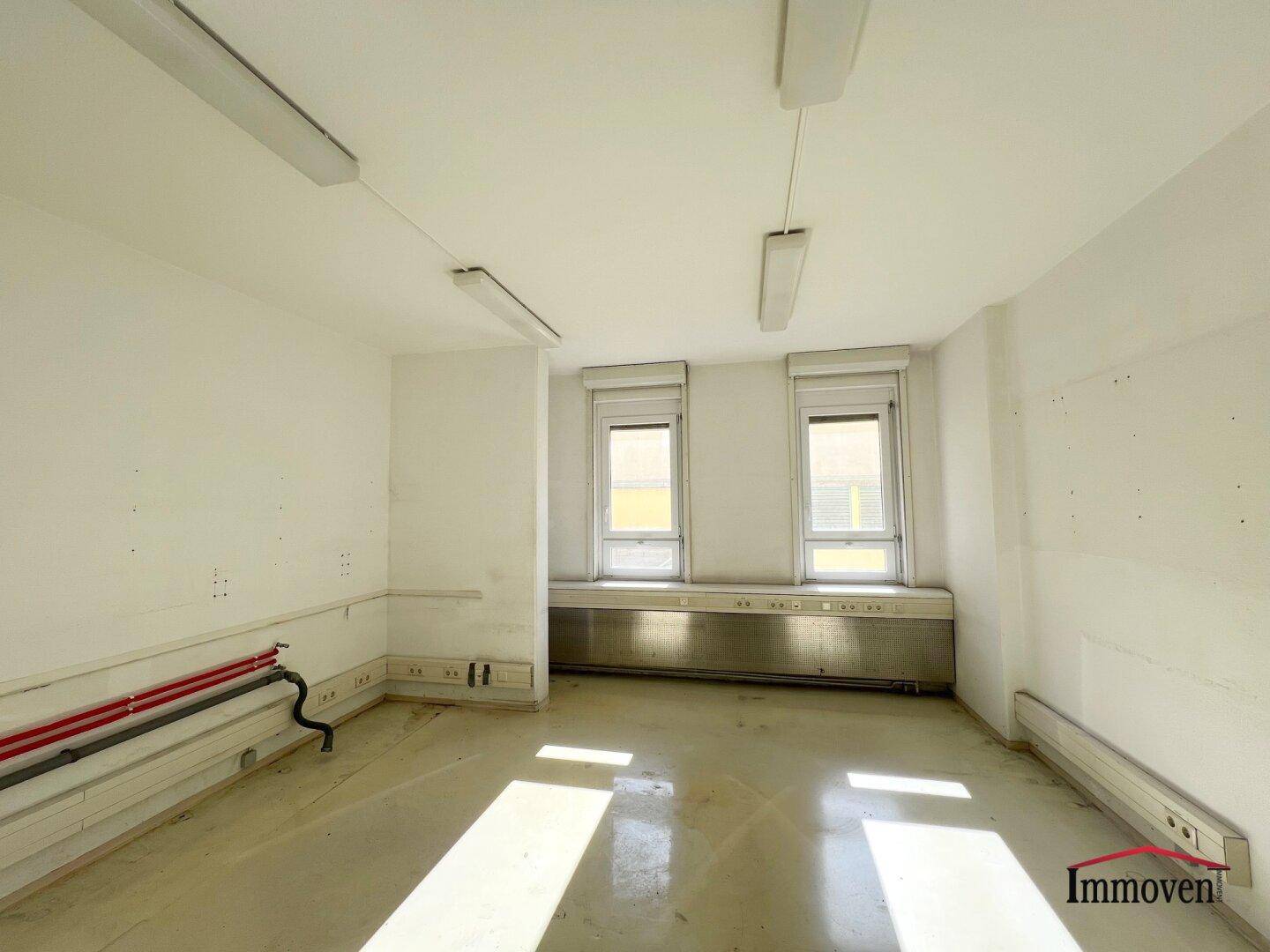 Geräumiger Büroraum mit zwei Fenstern, Jalousien und sichtbaren Leitungen an der Wand.