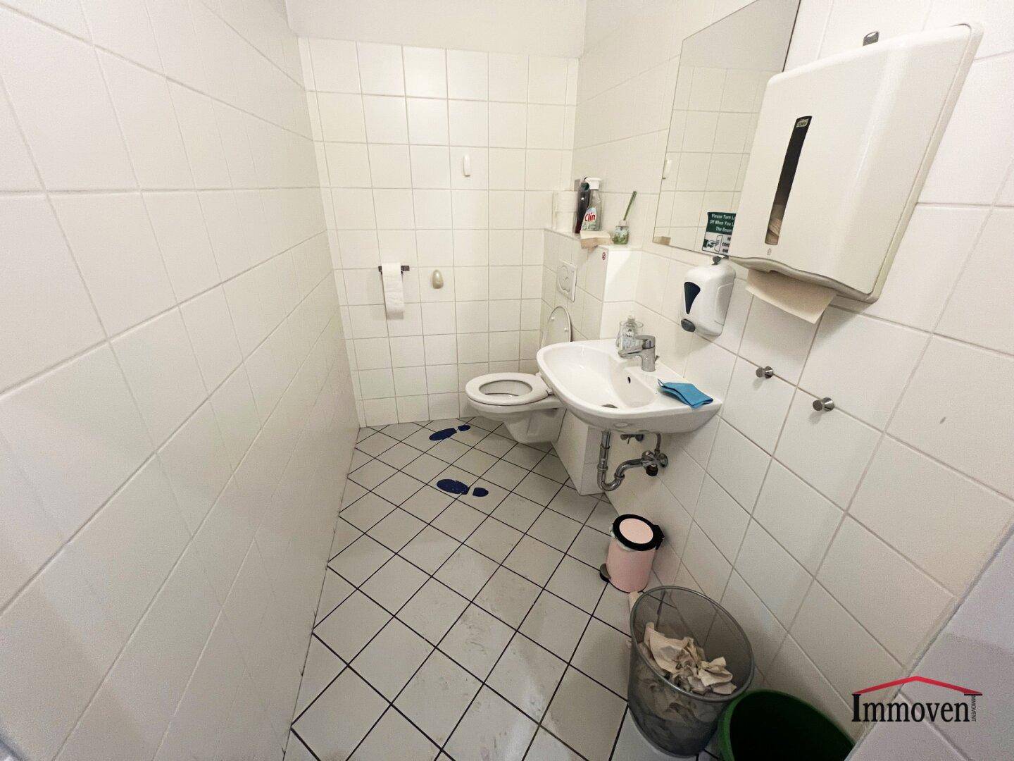 Toilettenraum mit weißen Fliesen, Waschbecken und Wand-WC, ausgestattet mit Spender.
