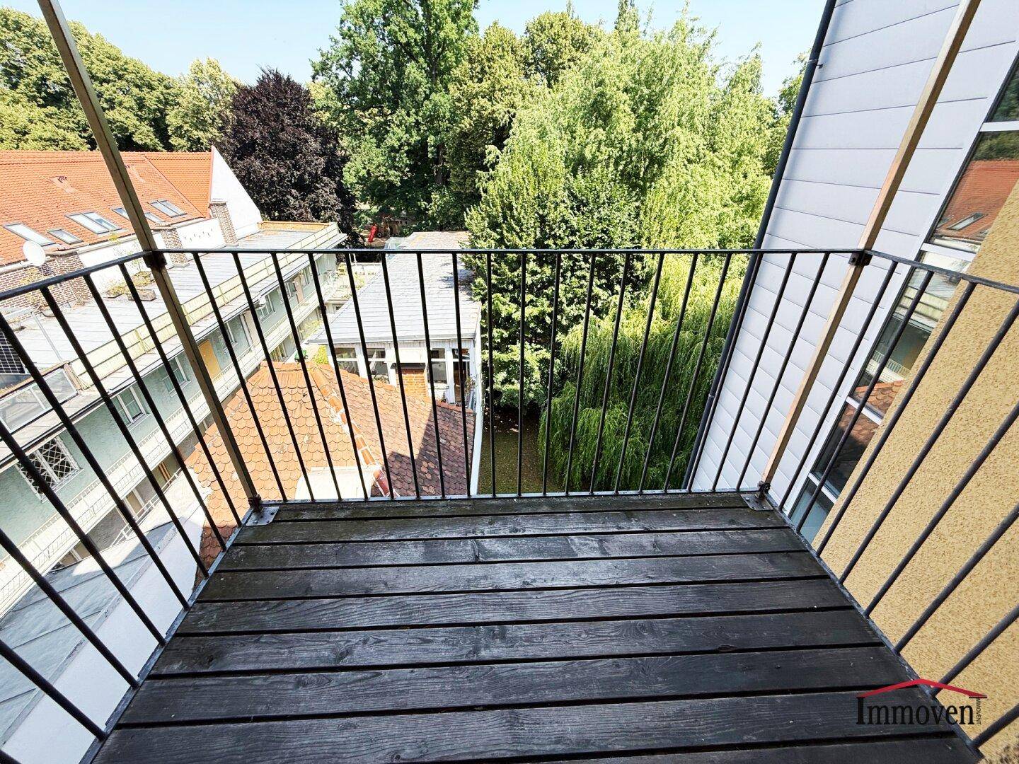 Kleiner Balkon mit Holzboden und Blick ins Grüne, umgeben von Bäumen und anderen Gebäuden.