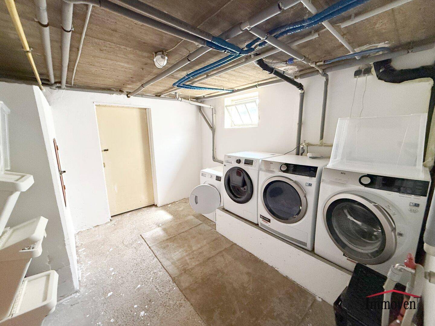 Waschküche im Keller mit Waschmaschinen, Trocknern und sichtbaren Leitungen.