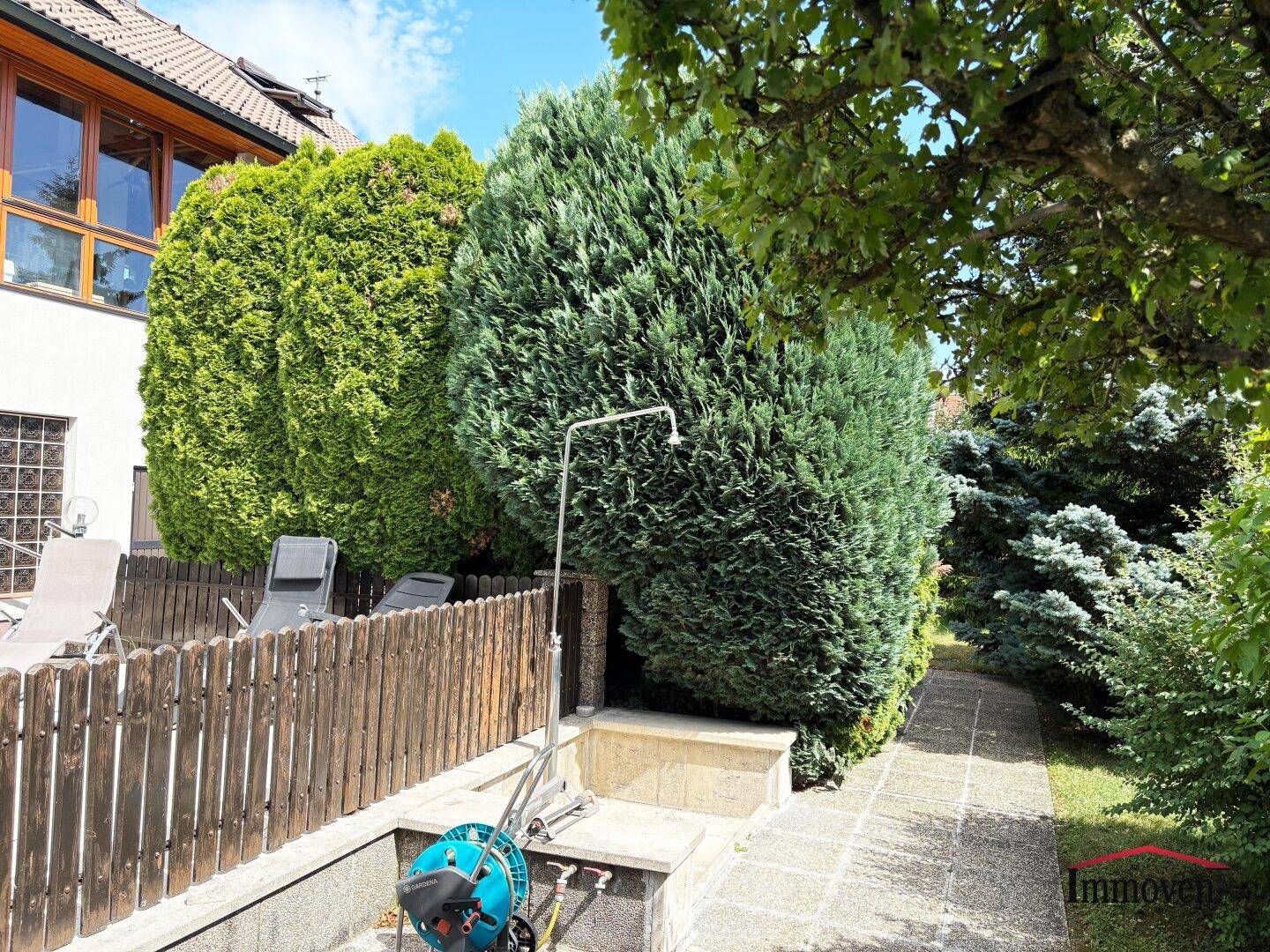 Gartenbereich mit hohen Hecken, Holzzaun und einer Außendusche für warme Tage.
