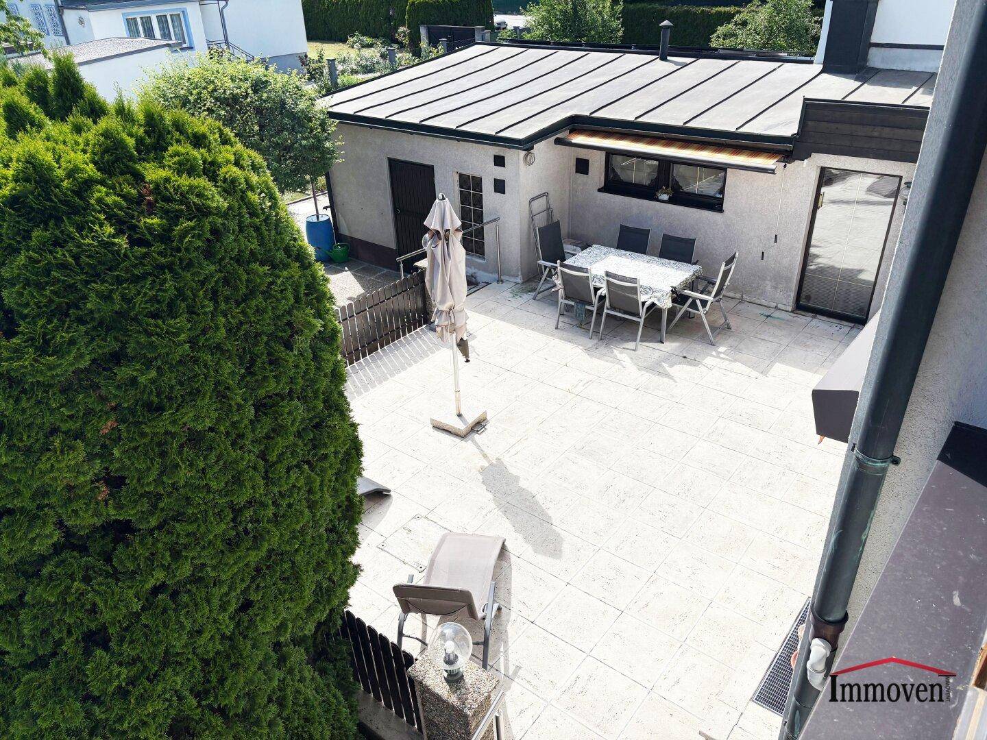 Große geflieste Terrasse mit Esstisch und Sonnenschirm, umgeben von Grün.