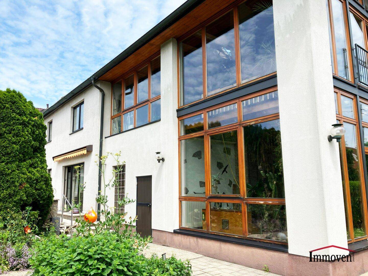 Moderne Hausfassade mit großen Glasflächen und Holzrahmen, umgeben von Grün.