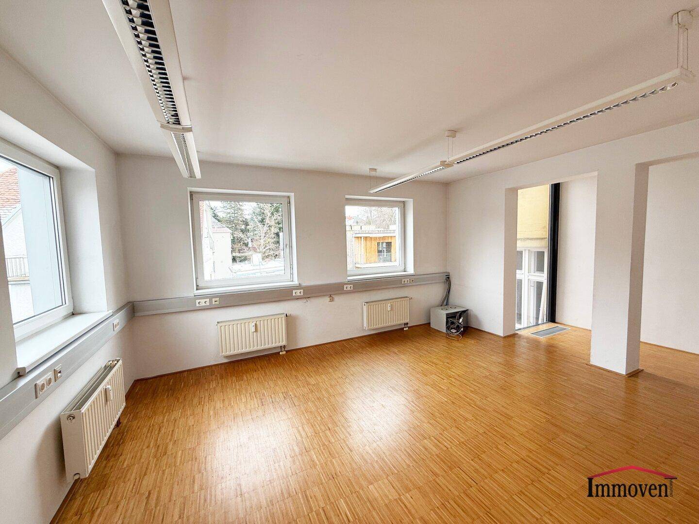 Heller Büroraum mit Holzparkett, großen Fenstern und Blick ins Grüne, ausgestattet mit Standardheizkörpern.