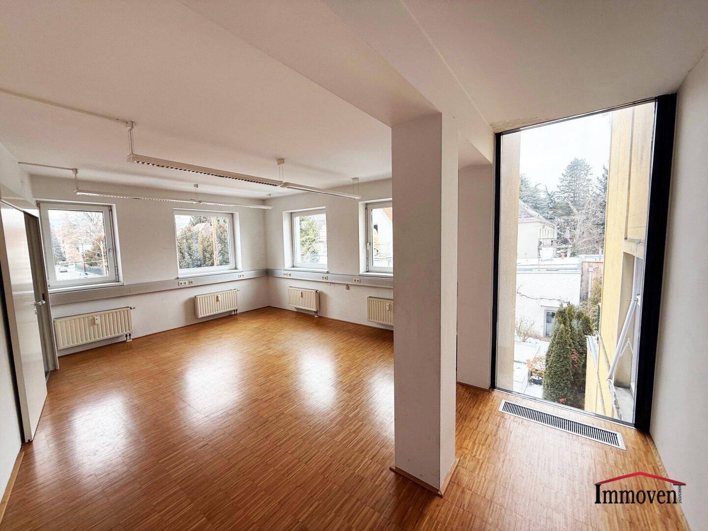 Geräumiger Büroraum mit Holzparkett, Säulen und einer großen Fensterfront, die viel Tageslicht hereinlässt.