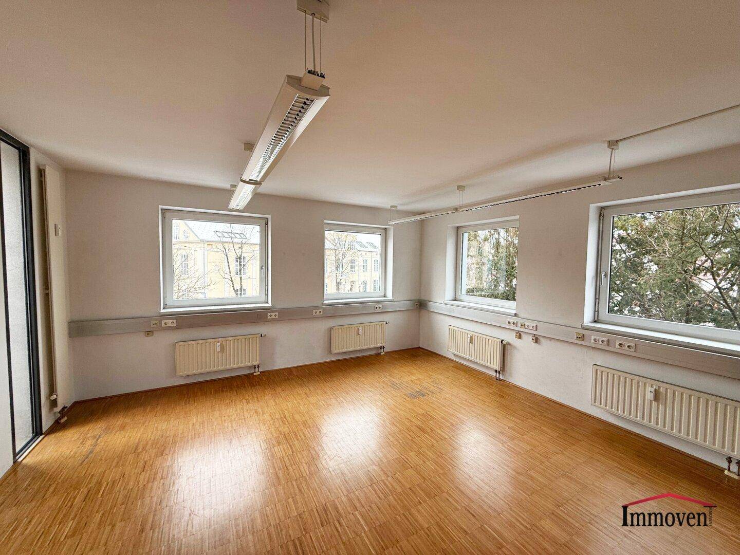 Leerer Büroraum mit Holzparkett, mehreren Fenstern und Deckenbeleuchtung, ideal für flexible Gestaltung.