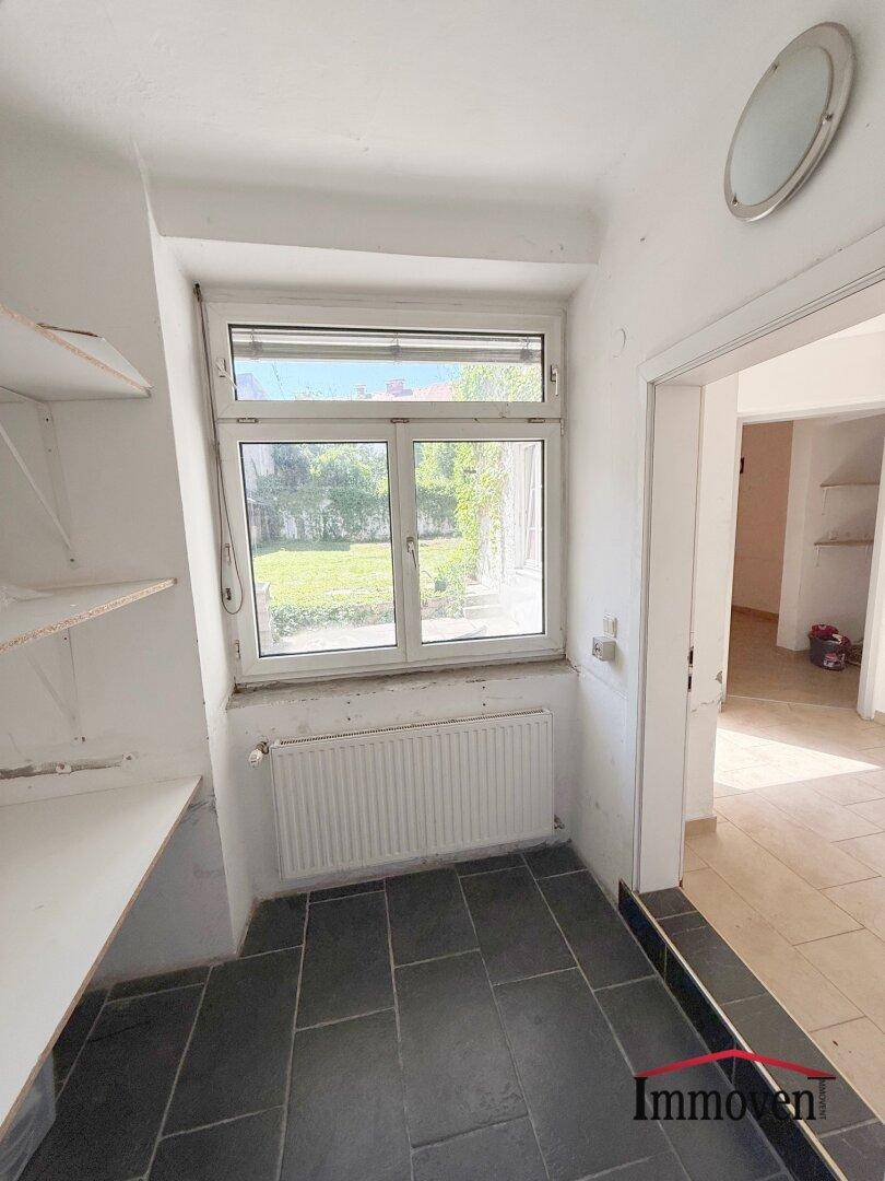Abstellraum mit Fenster und Regalen, der zu einem weiteren Raum mit hellem Fliesenboden führt.