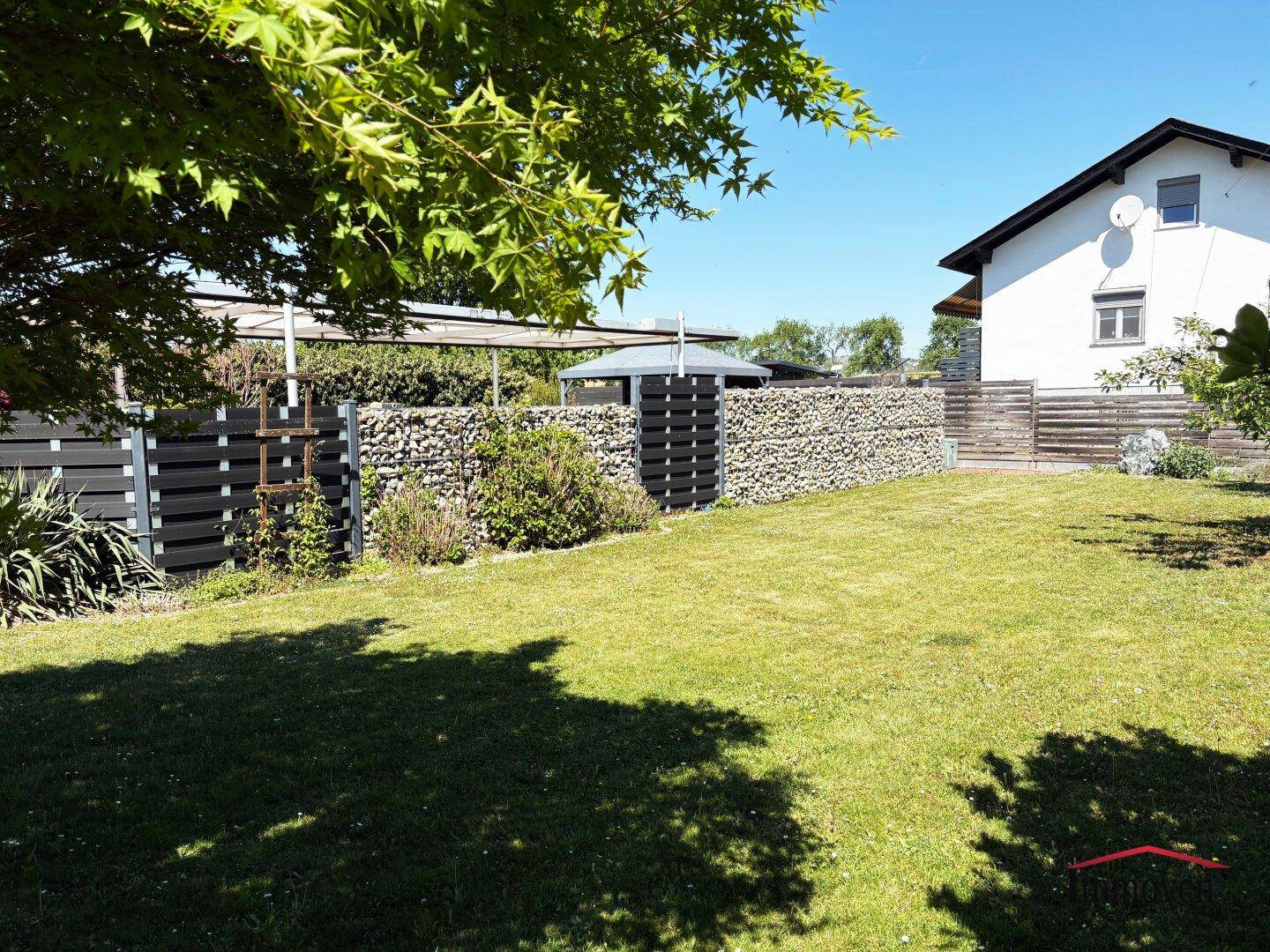 Großer Garten mit Rasenfläche, Gabionenwand und Blick auf das Wohnhaus unter blauem Himmel.