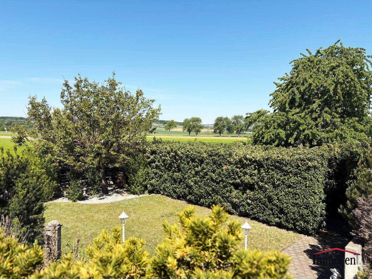 Panoramablick vom Garten über die grüne Hecke auf die weite, offene Landschaft unter blauem Himmel.