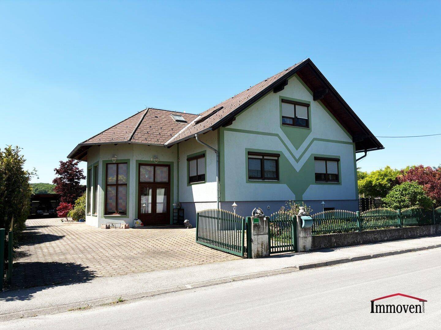 Freistehendes Haus mit Satteldach, gepflasterter Zufahrt und grünem Zaun an der Straße.