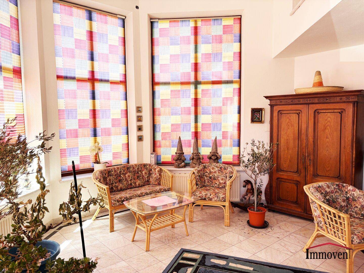 Gemütlicher Sitzbereich im hellen Wintergarten mit Korbmöbeln und bunten Fensterrollos.