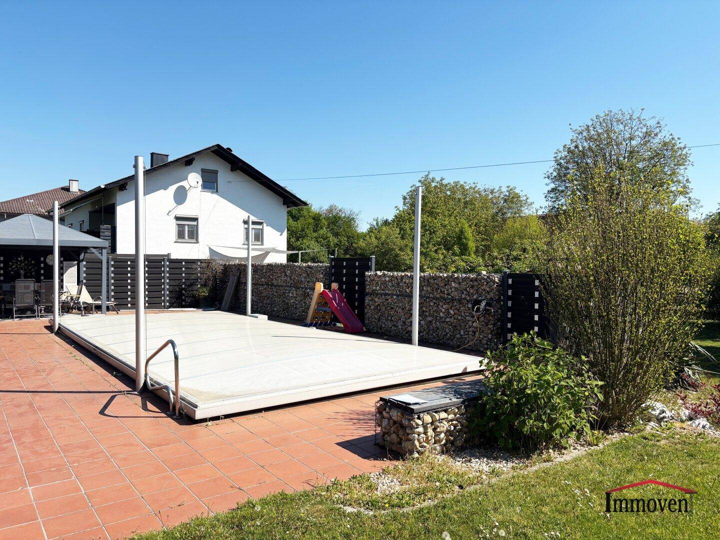 Außenbereich mit abgedecktem Pool, Terrasse und einem Haus im Hintergrund unter klarem Himmel.