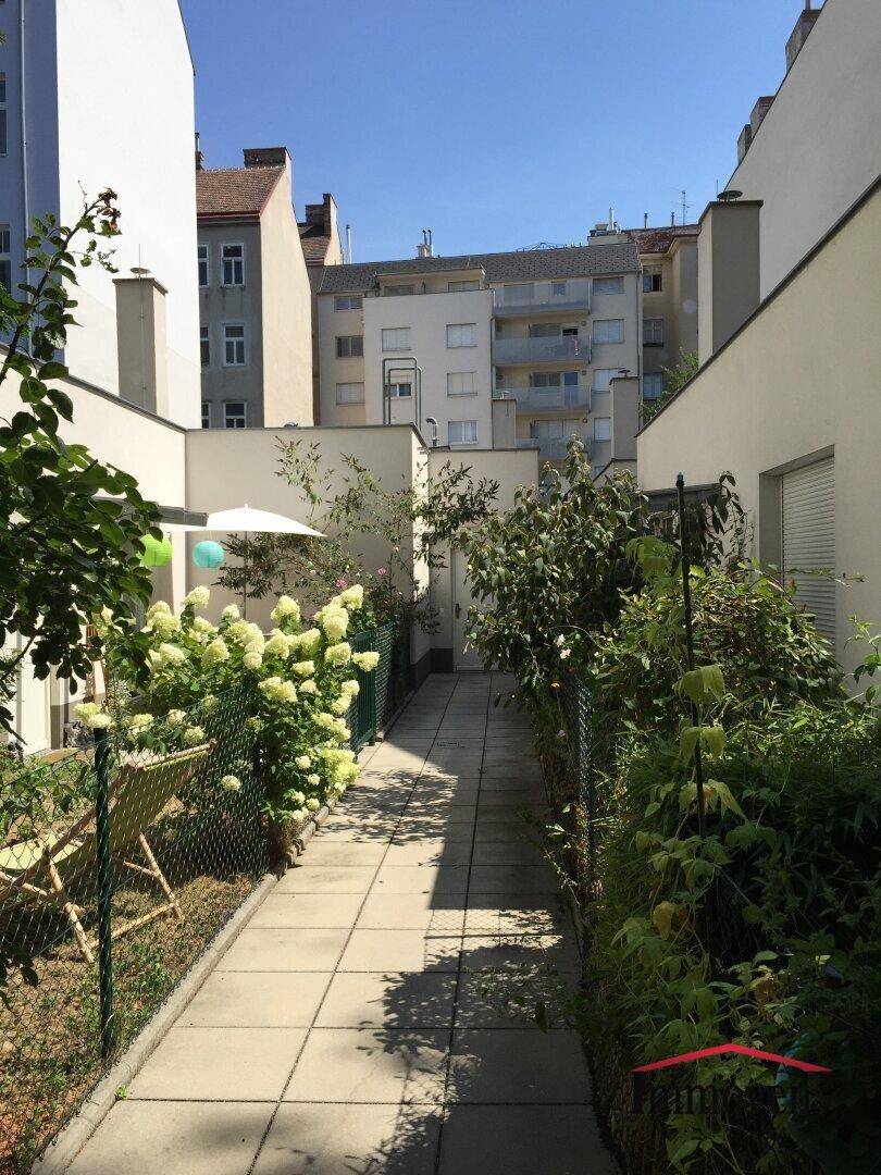 Gepflegter Gartenweg zwischen Gebäuden mit üppiger Bepflanzung und Sitzgelegenheiten im Freien.
