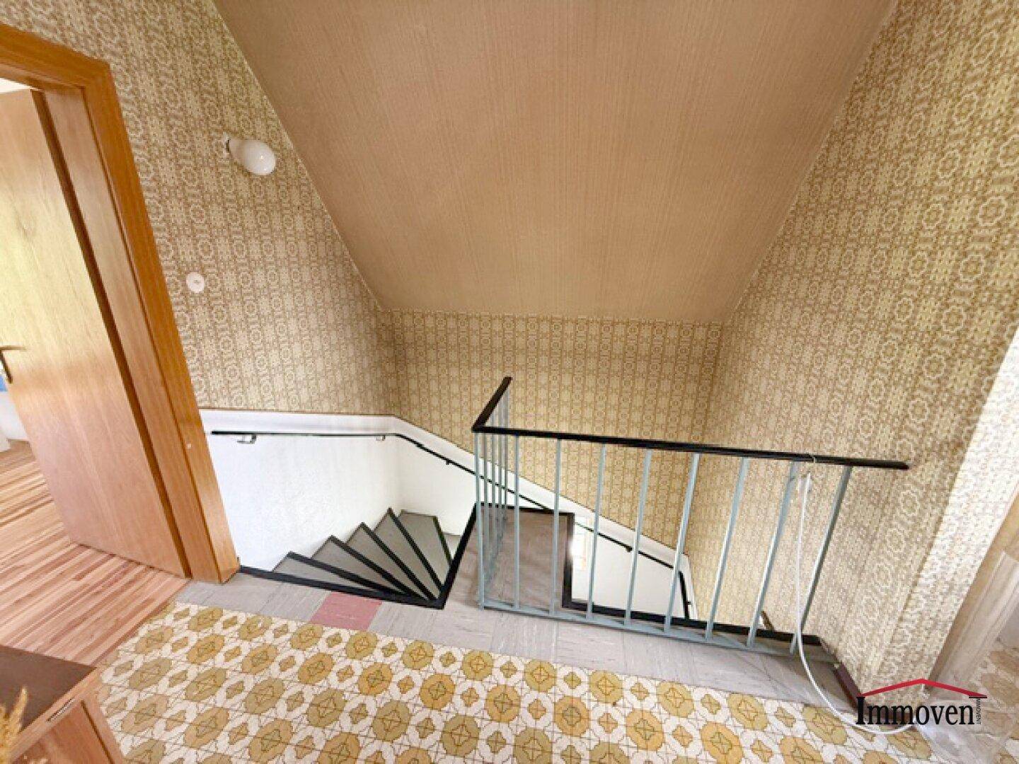 Treppenhaus mit gemusterter Tapete und Holzgeländer, das in die obere Etage führt.