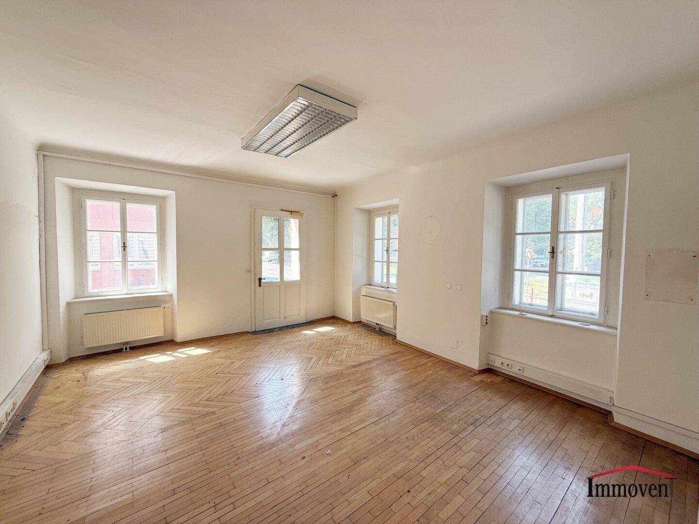 Helles Zimmer mit Holzboden, hohen Decken und mehreren Fenstern, ideal für Wohn- oder Büronutzung.