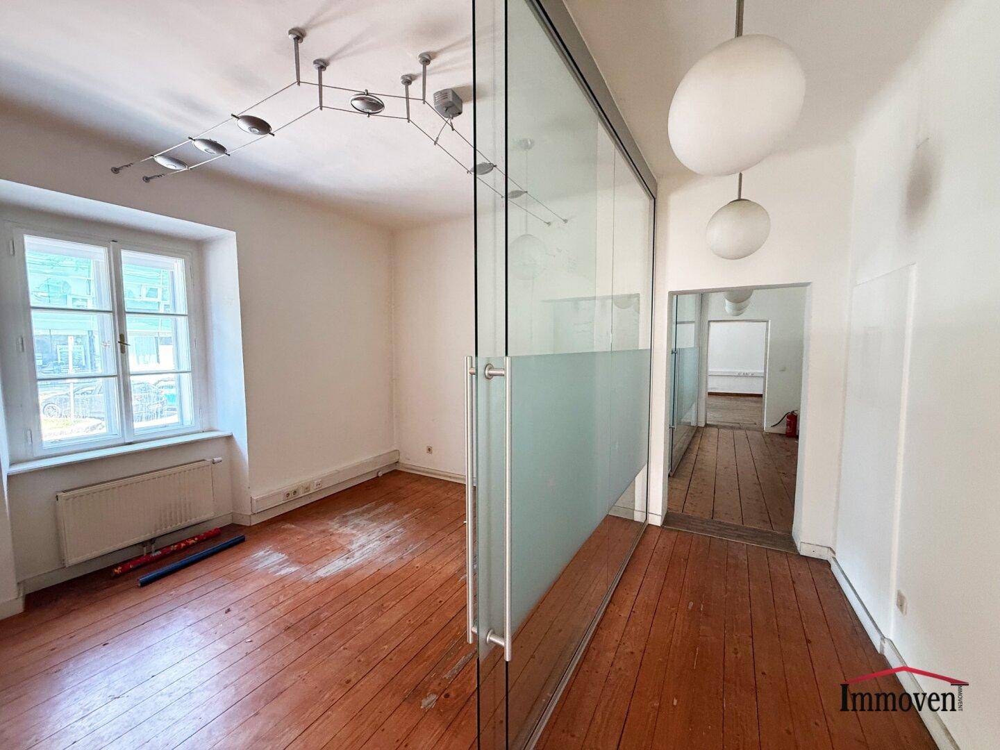 Modernes Büro mit Glaswand, Holzboden und Deckenleuchten, das eine helle Arbeitsumgebung bietet.