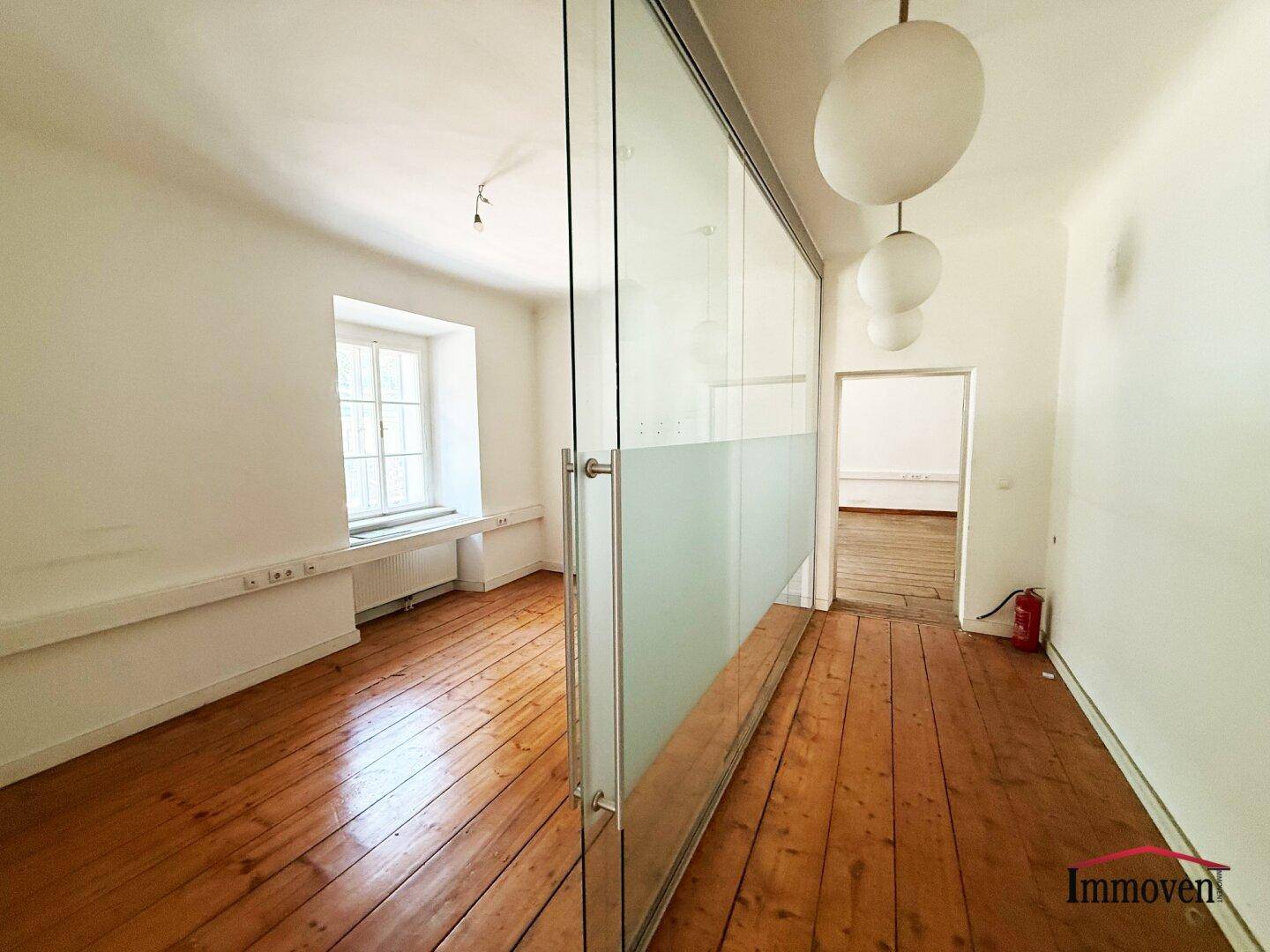 Heller Flur oder Bürobereich mit Holzboden, Glaswand und modernen Hängelampen, die den Raum erhellen.