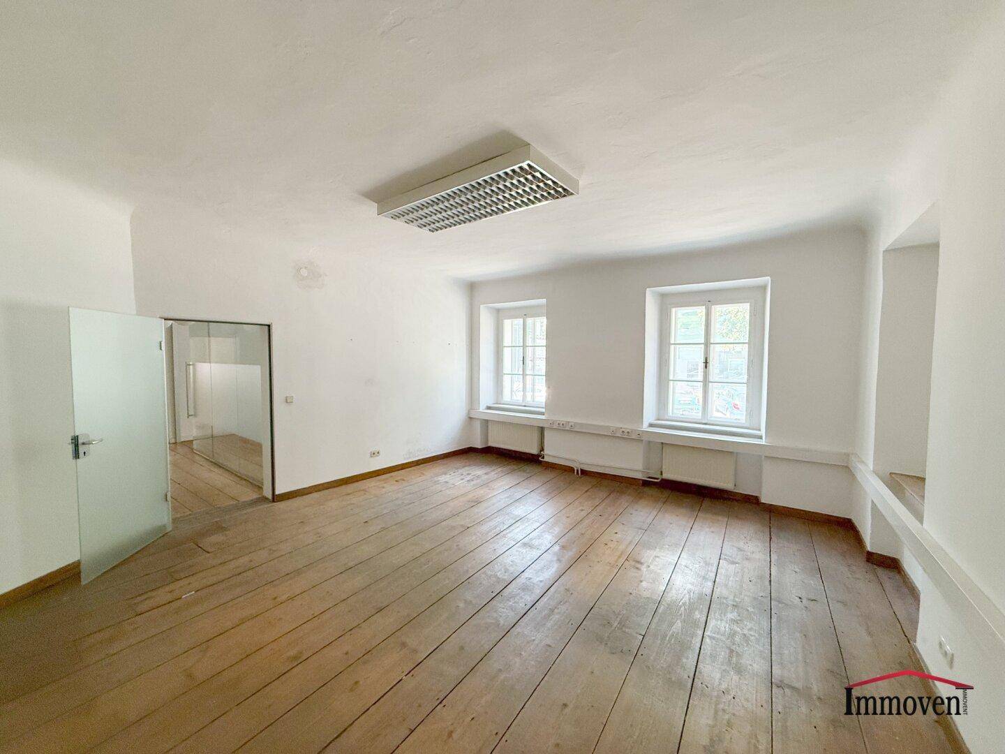 Großes, helles Zimmer mit Holzboden und zwei Fenstern, das sich gut als Büro oder Arbeitsraum eignet.