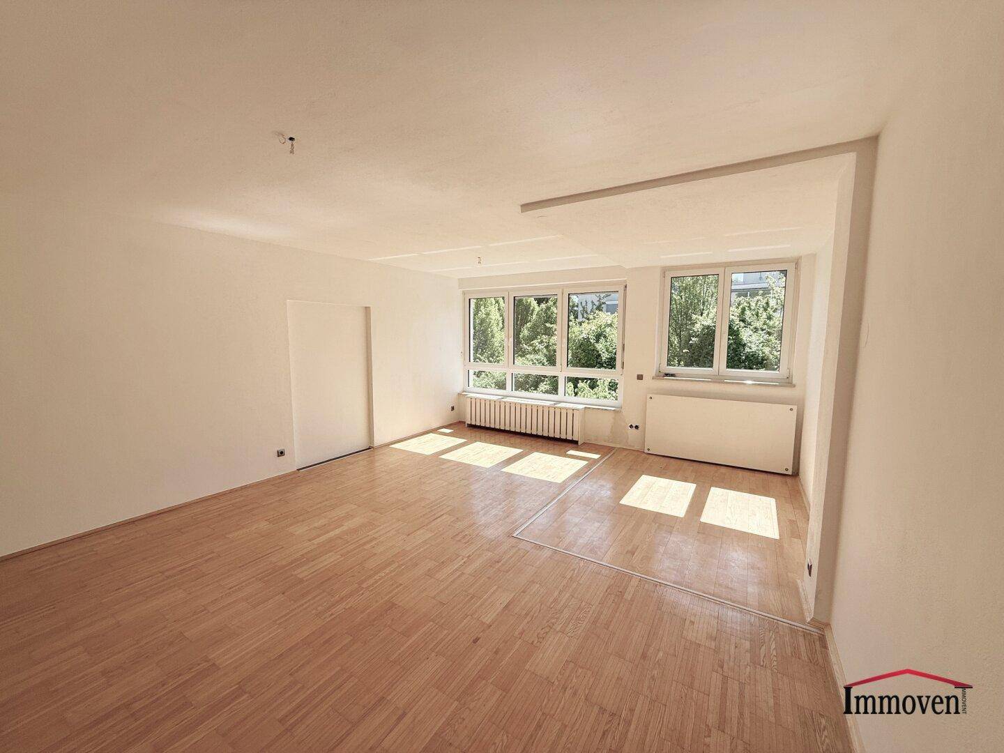 Geräumiges Wohnzimmer mit viel Tageslicht, hellem Parkettboden und weißen Wänden.