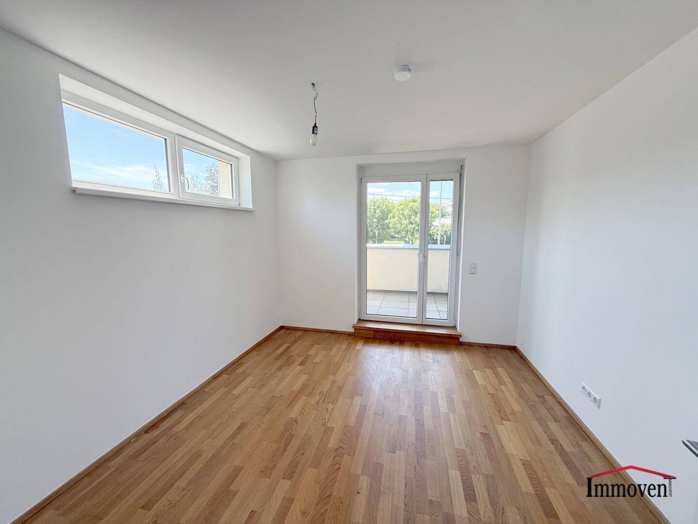 Helles Zimmer mit Holzboden, einem Fenster und Zugang zu einem Balkon.