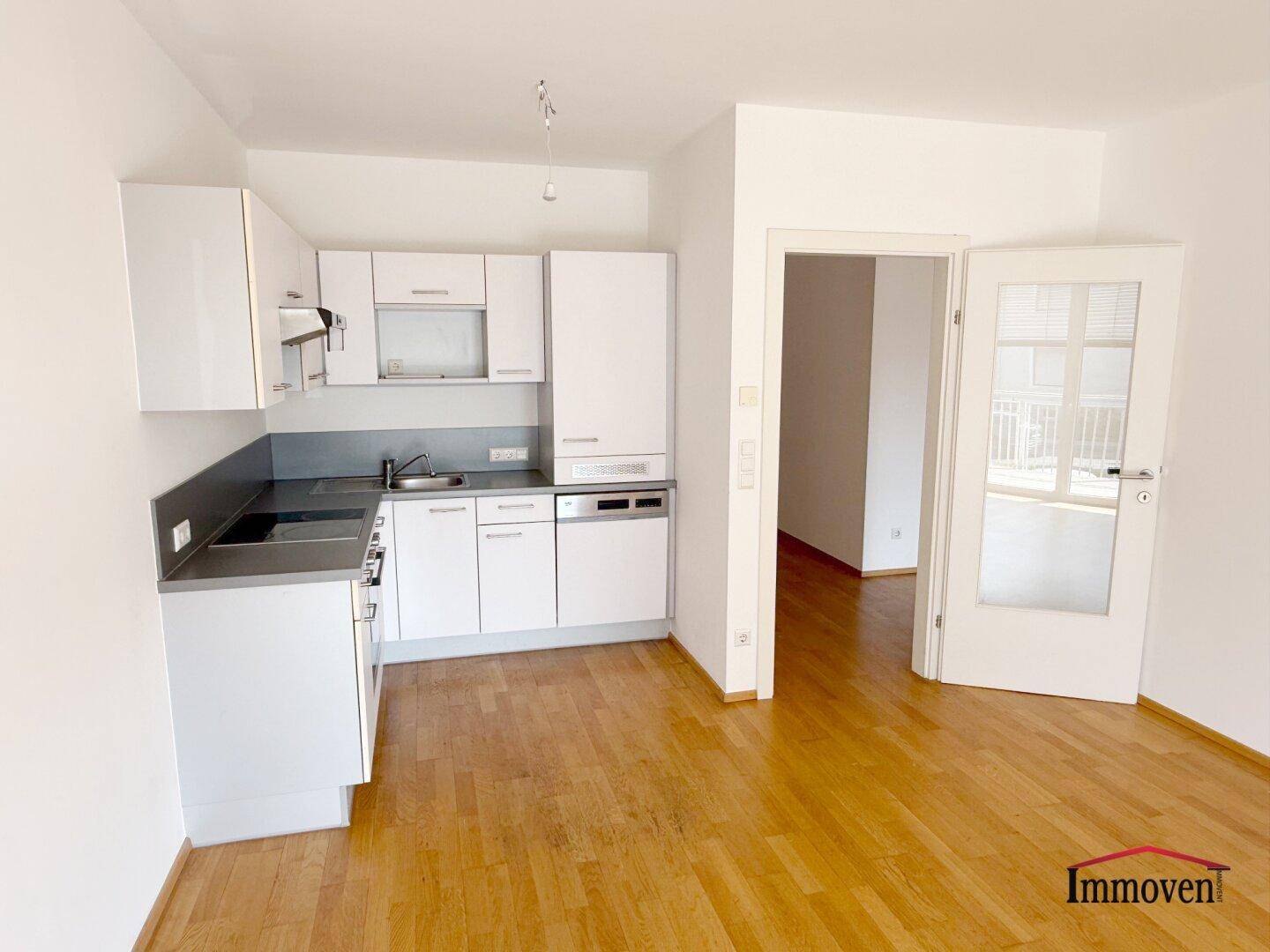 Moderne Einbauküche mit weißen Schränken, grauer Arbeitsplatte und Parkettboden in offenem Bereich.