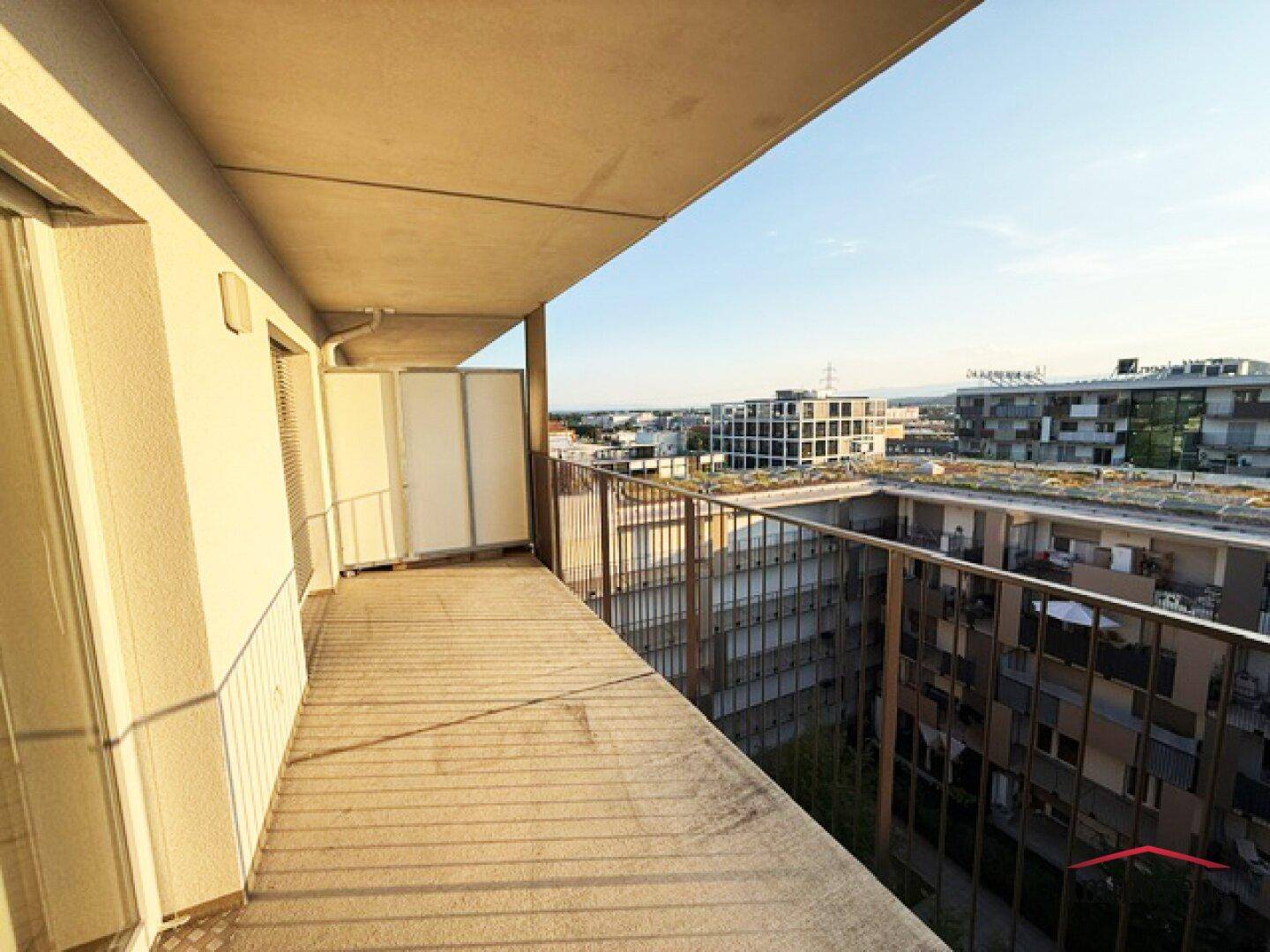 Langer Balkon mit Metallgeländer und Blick auf die umliegenden Wohngebäude bei Sonnenschein.
