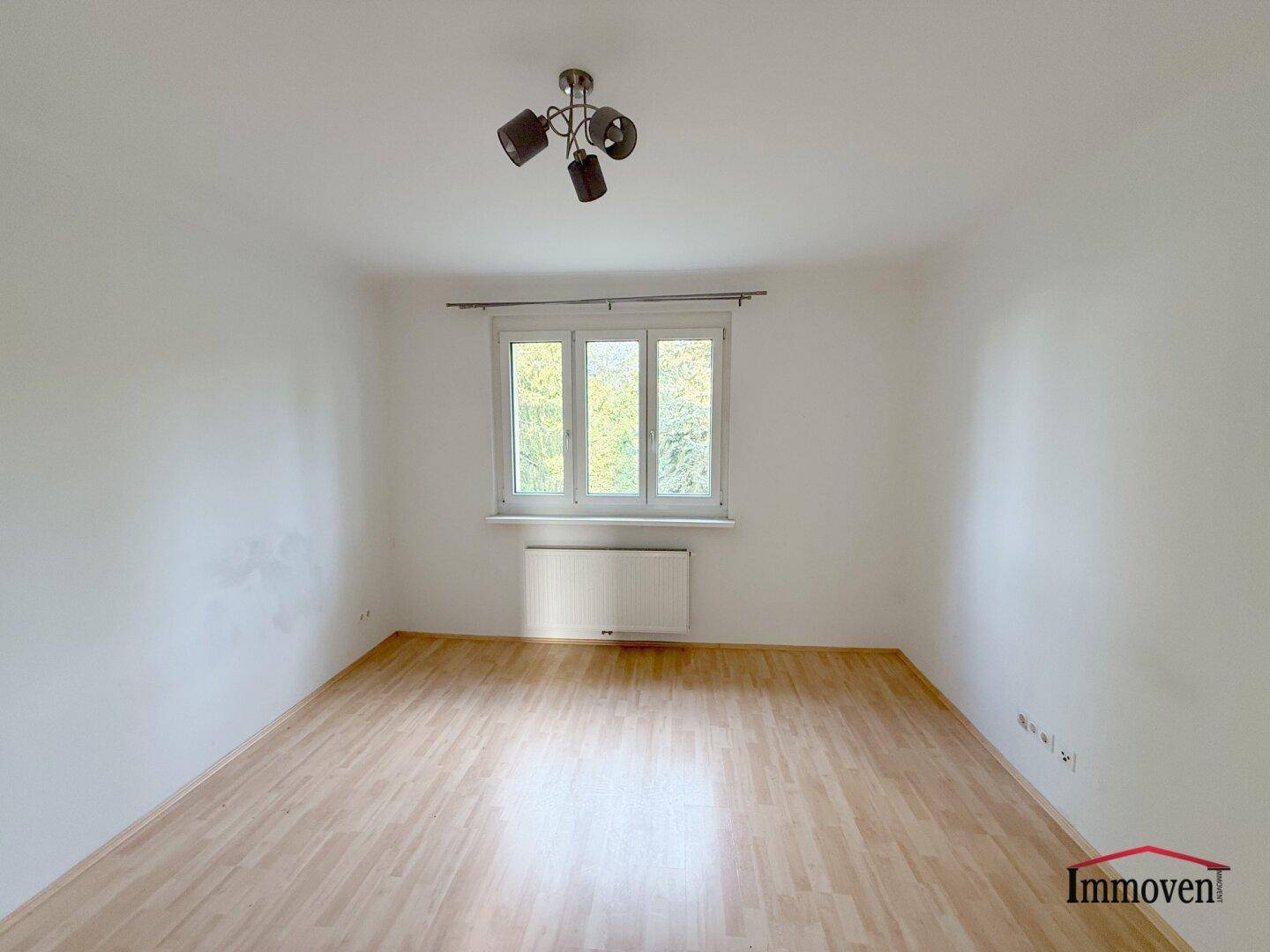 Gemütliches Zimmer mit einem Fenster, hellem Laminatboden und schlichter Deckenleuchte.