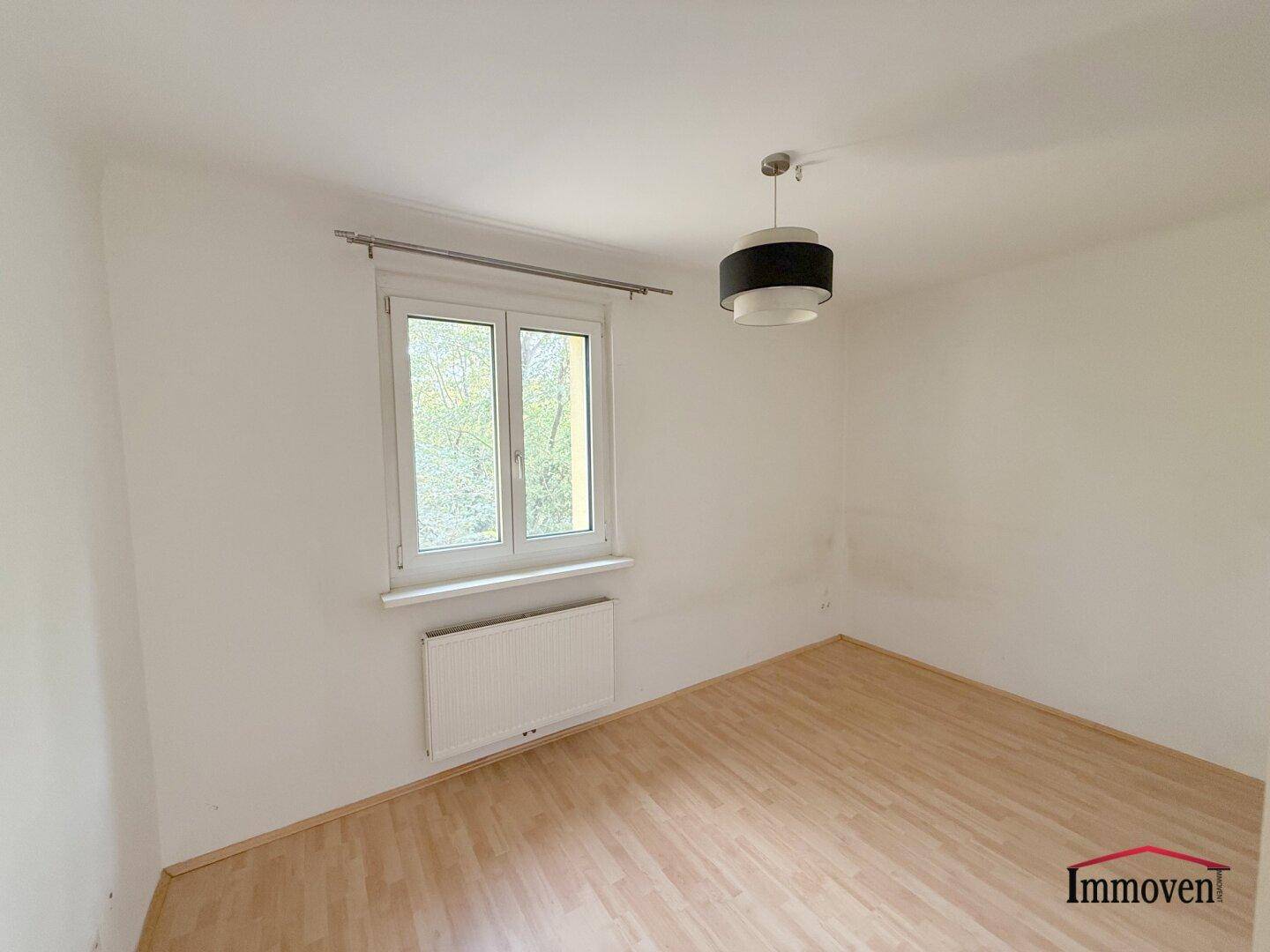 Freundliches Zimmer mit Laminatboden, einem Fenster und moderner Deckenlampe.