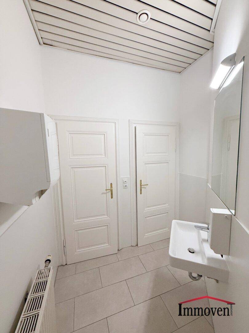 Toilettenraum mit Waschbecken, zwei Türen und einer Holzlamellendecke, funktional eingerichtet.